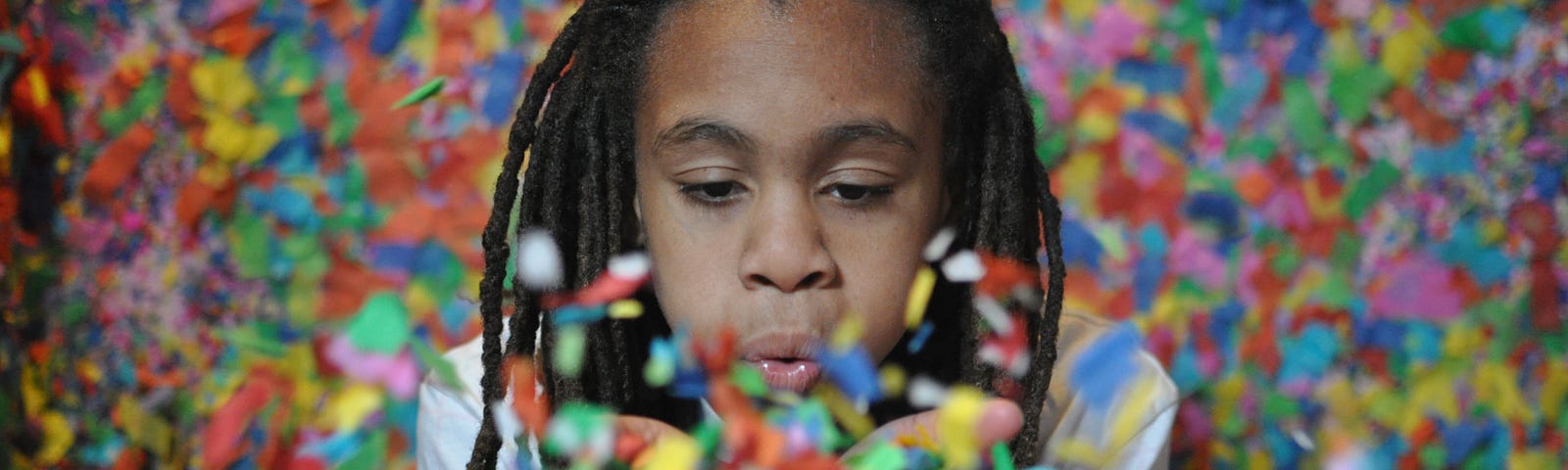 In front of a wall of confetti, a girl blows confetti from her hands to celebrate.