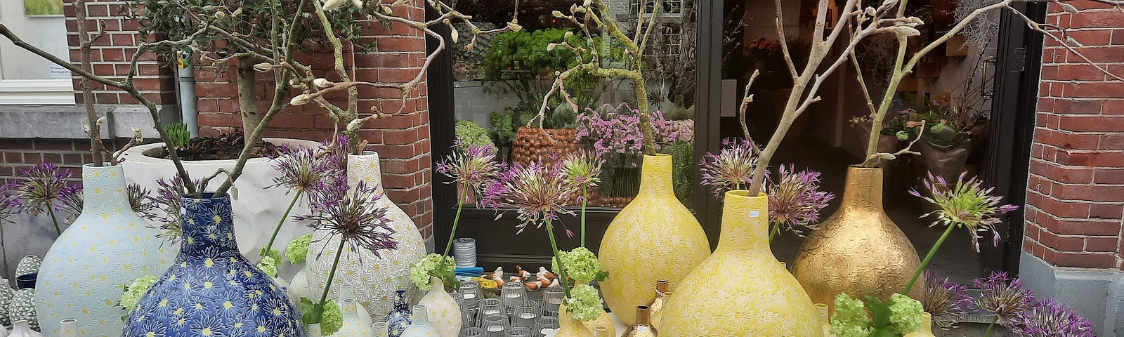 Two white tables containig vibrant ywllo, blue, and white colored vases of different sizes holding flowers and Magnolia branches.