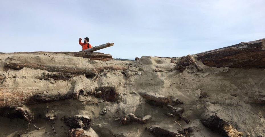 Man in a bright orange sweater standing on top of a cliff of driftwood and sand and looking out into the distance, his hand shading his eyes from the sun