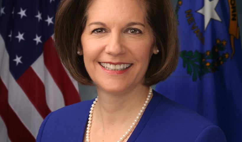 Nevada Senator Cathetrine Cortez Masto’s official portrait. She is wearing a royal blue suit jacket and pearls, and she is in front of an American flag and the Nevada state flag.