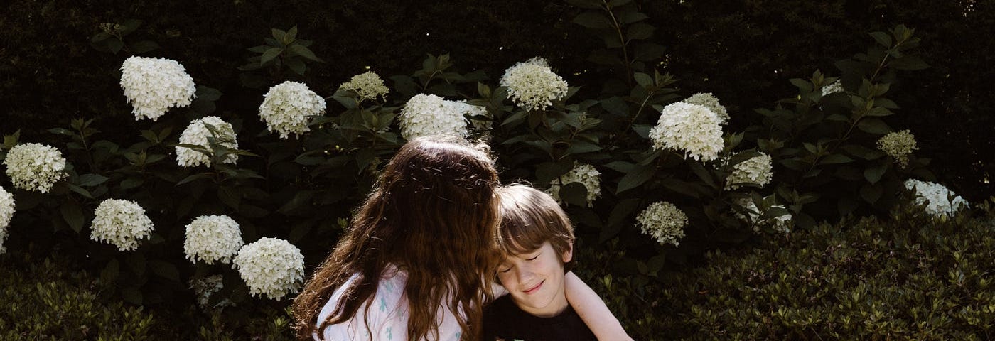 A school-age sister hugs her little brother as they sit together on a bench in front of white hydrangeas.