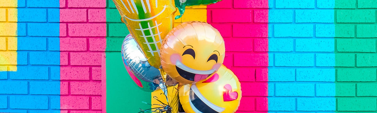 A woman standing in front of a colorful wall. Her face is covered by air baloons shaped like emoticons and an ice cream cone.