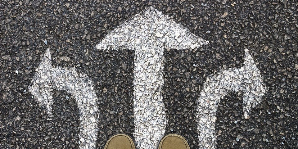 A view looking down at two feet in brown shoes standing on concrete with three white arrows on it. On arrow points left, another straight ahead and the third to the right.