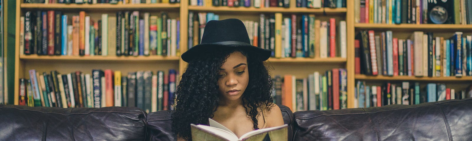 girl wearing a hat and reading a book on a couch, with bookshelves in the background