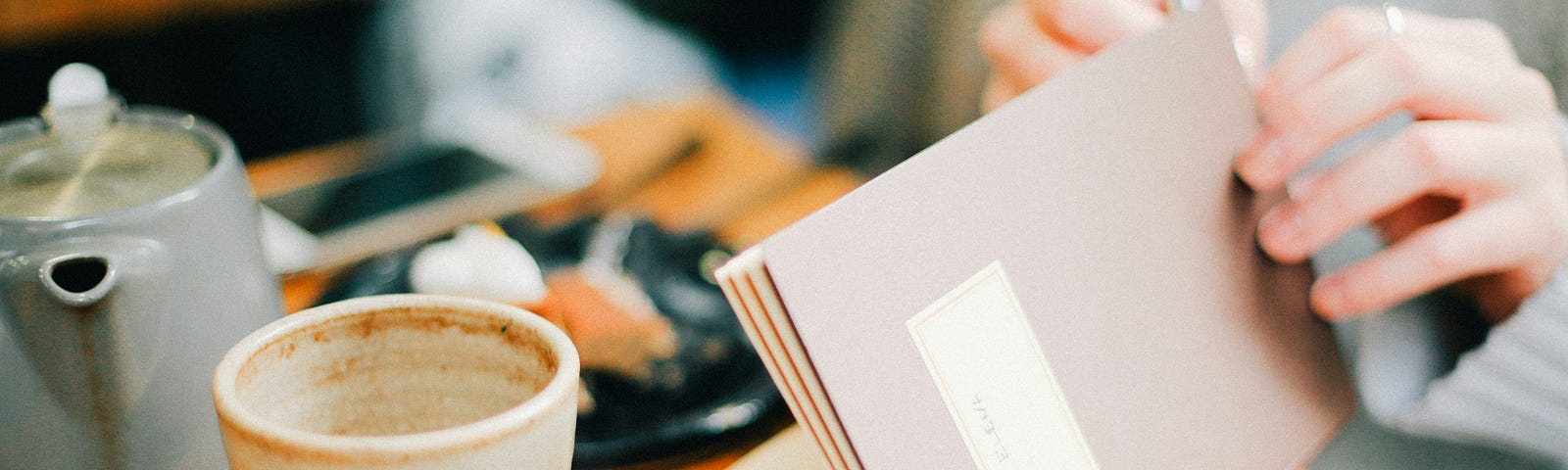 A person writing in a notebook with a cup of tea and a teapot on the table, creating a serene and reflective atmosphere.