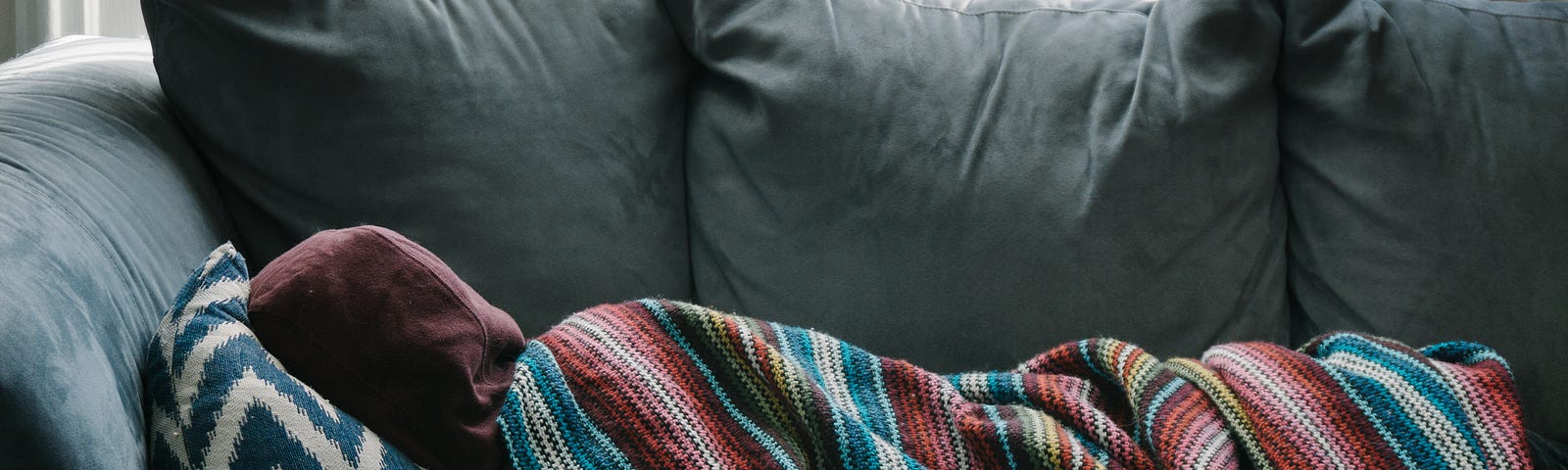 A person lying on a couch, wearing a hoodie and using a pillow and blanket.