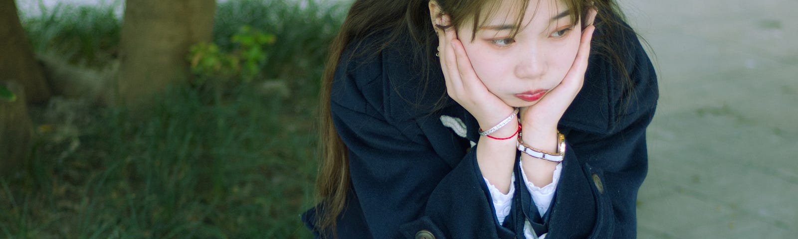 A teenage girl sits head in her hands, dressed in a blue school uniform of blue dress, blue blazer, black socks and mary janes.