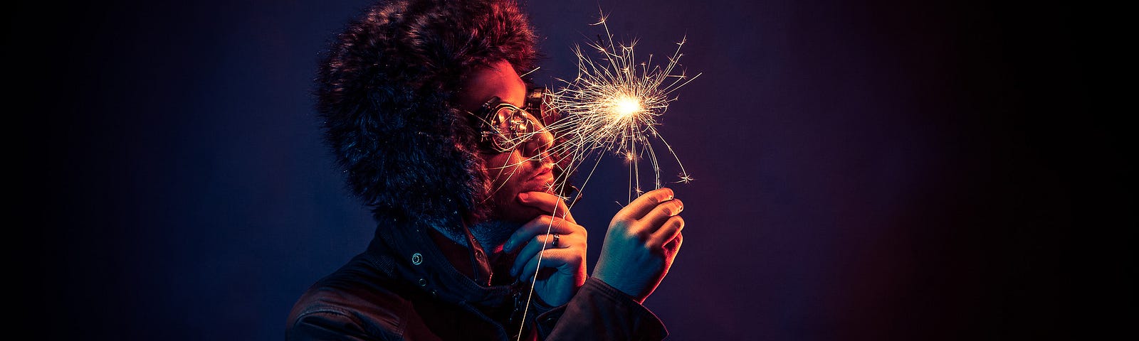 dark photo of a man wearing a jacket, a fuzzy hat and goggles while holding a sparkler in front of his face.