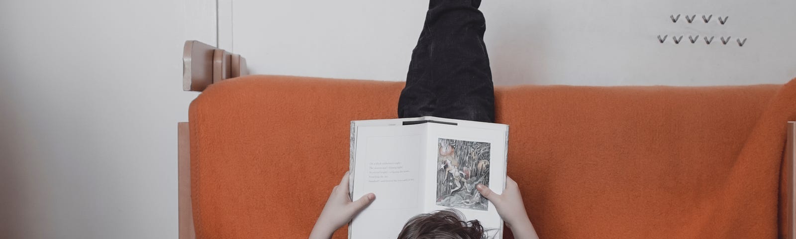 Someone is laying on the ground with their feet up on a couch. They are reading a book. Behind their head on the ground is a huge stack of more books.