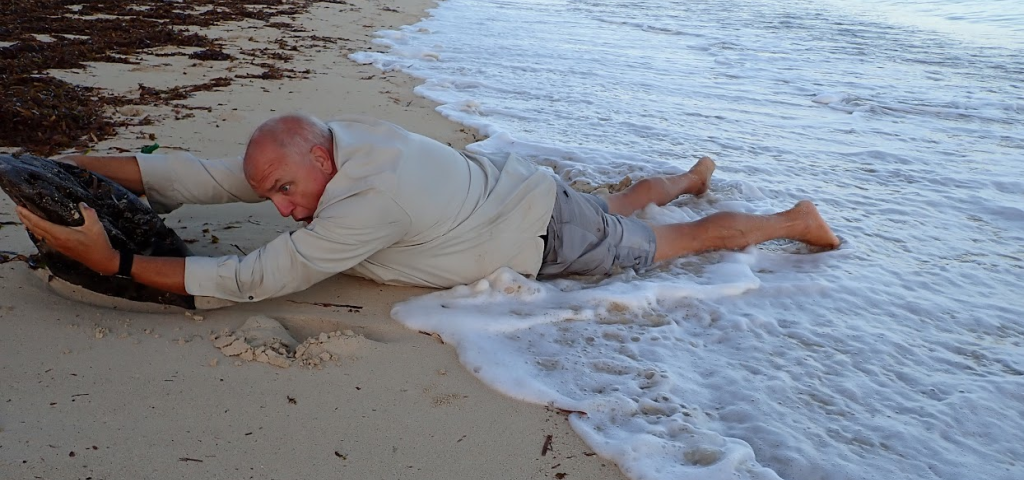 Author’s husband having some fun- lying face-down on the beach in the surf, holding onto a log. He has a scared look on his face like he is holding on for dear life not to be washed out to Sea.