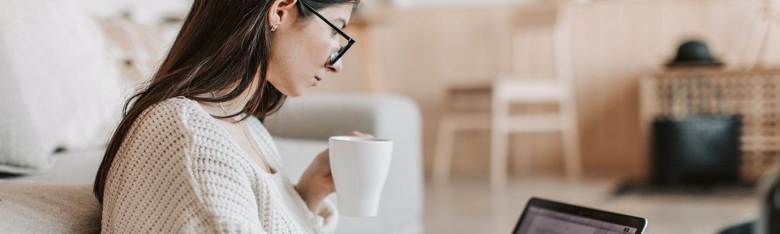 a girl sitting on floor in white sweater reading something on a laptop while drinking coffee in a white mug.