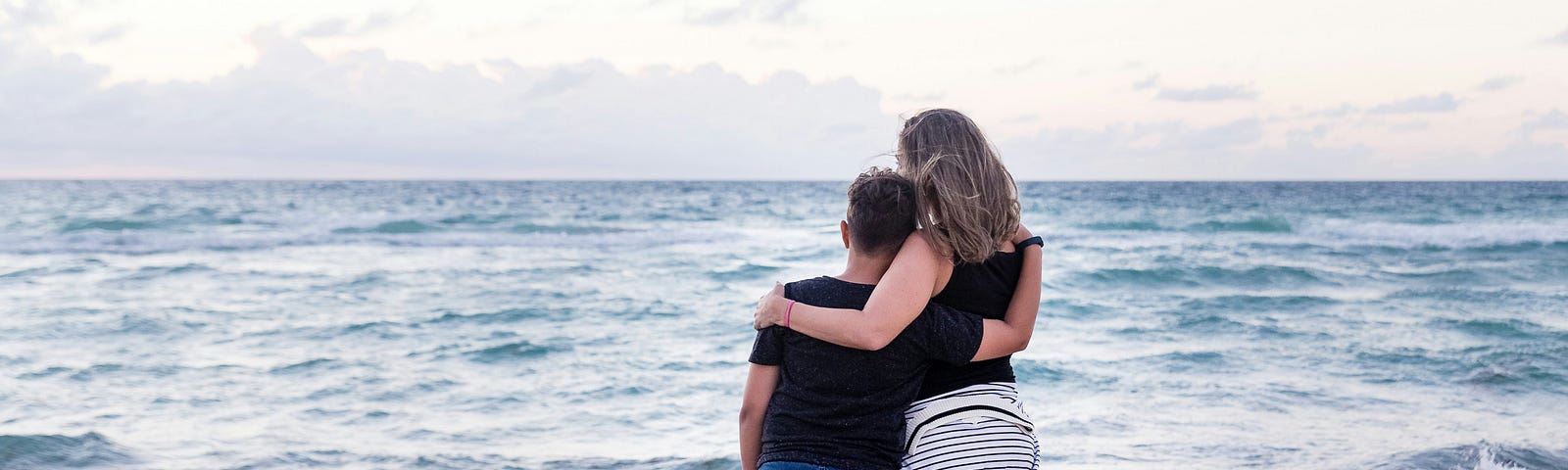 An image of a mother and son hugging standing on a beach and looking out to sea