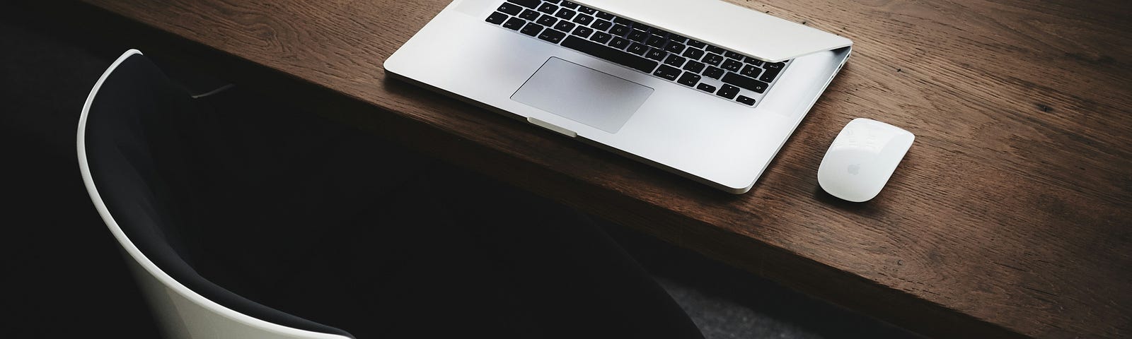 Dark wood desk with laptop half closed, mouse, and modern white chair.