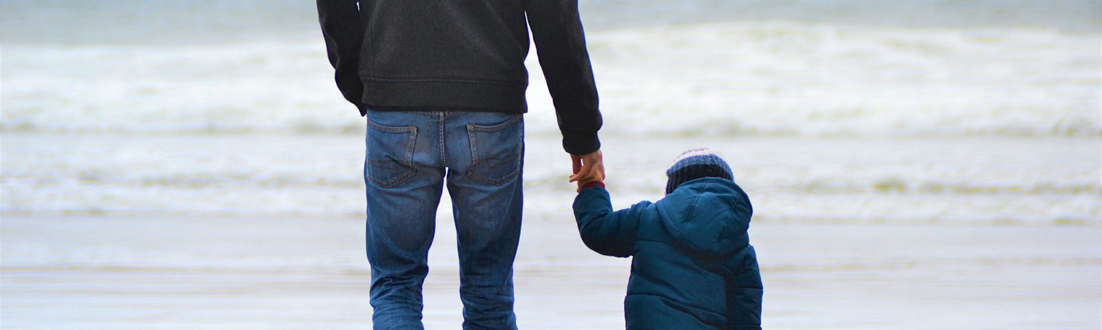 a dad walking hand-in-hand with his son along a beach with waves crashing the shoreline.
