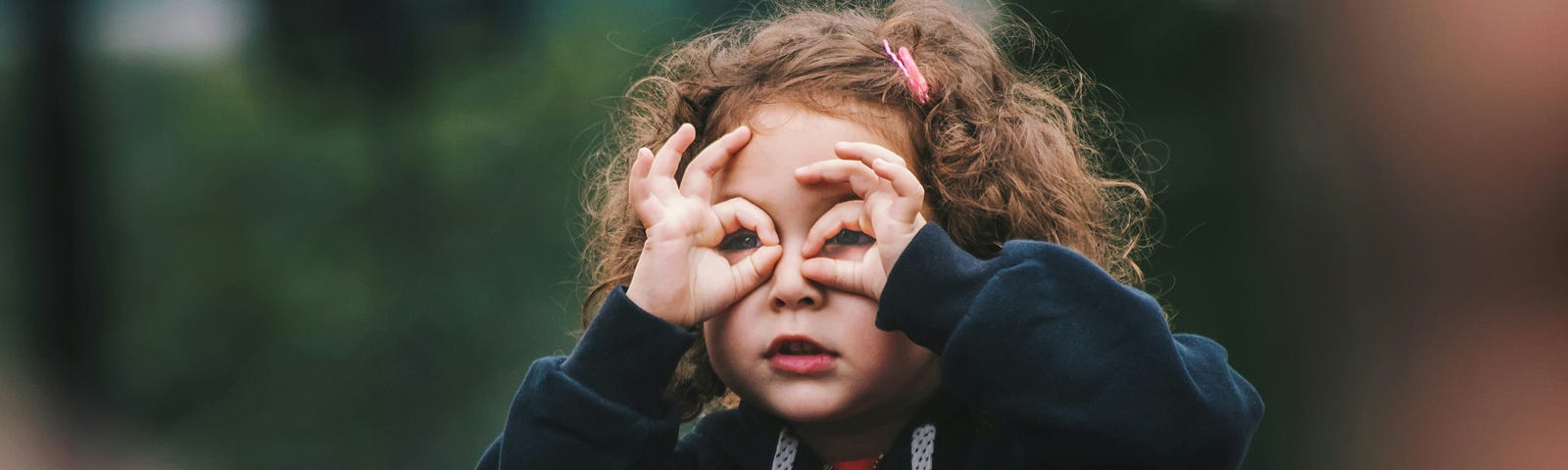 Little girl on her daddy’s shoulders with her fingers cupped around her eyes, mimicking eyeglasses.