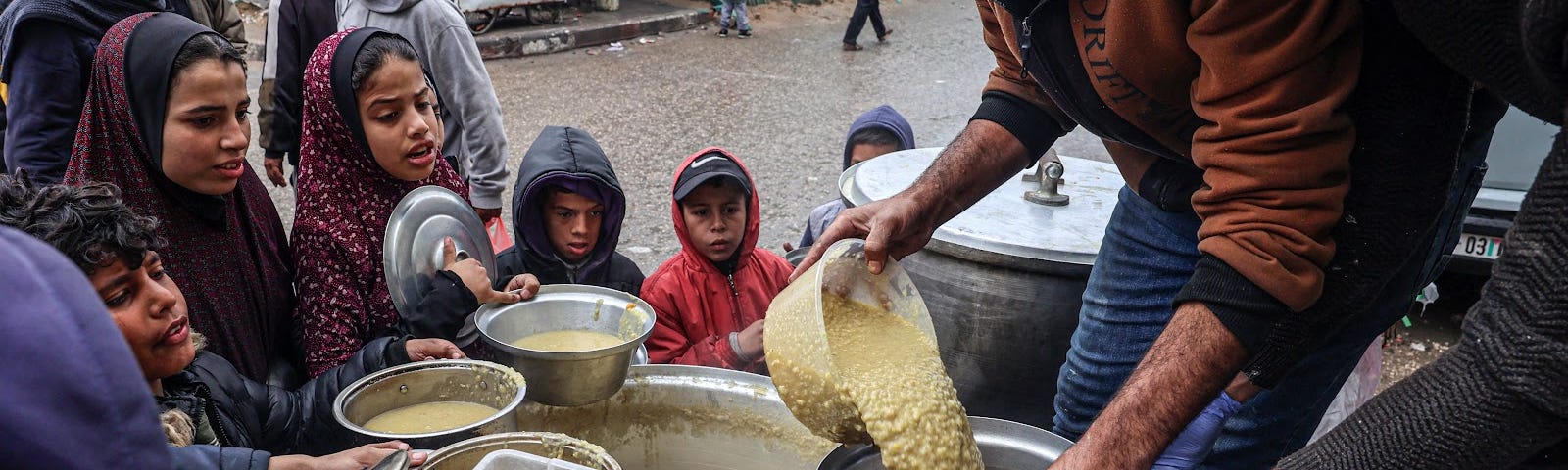 Two men pour lentils into a pot from a large vat while children circle them with their containers outreached, waiting for their share.
