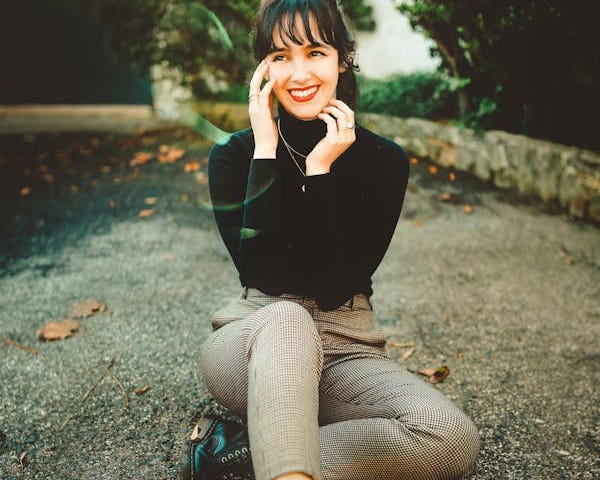 Young woman sitting on asphalt road with autumn trees out of focus in the background. She’s looking away from the camera with her hands to her face, smiling very wide