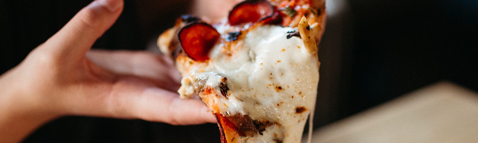 A hand holding a slice of pizza over a pie, the cheese and toppings sliding off in strings.