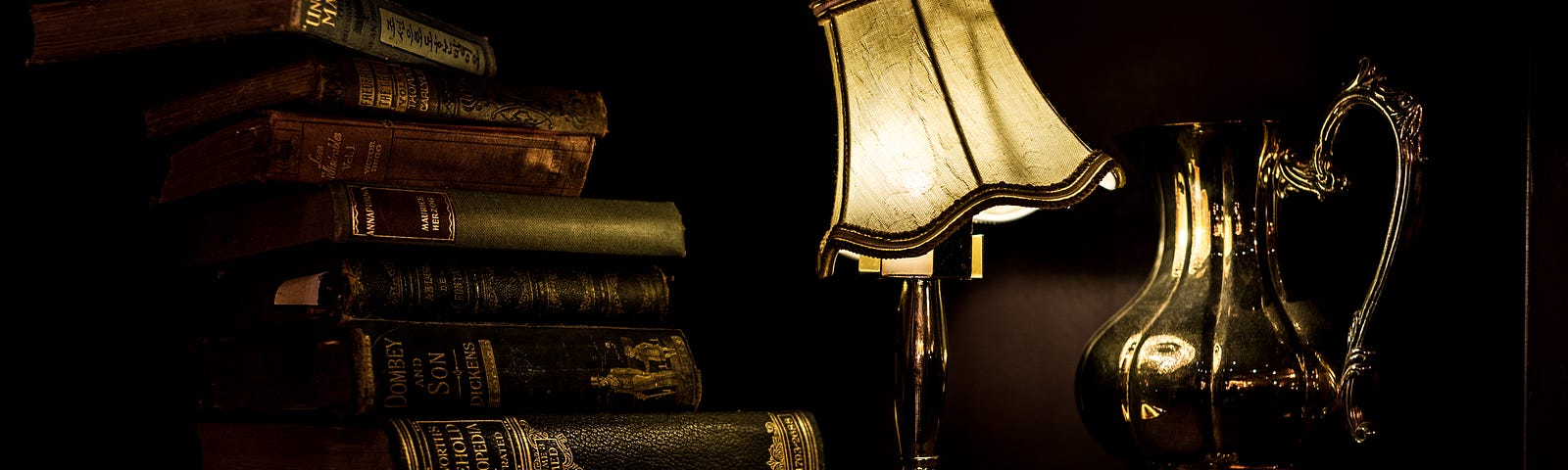 a lamp with a tilted shade between a silver pitcher and a stack of hardback books