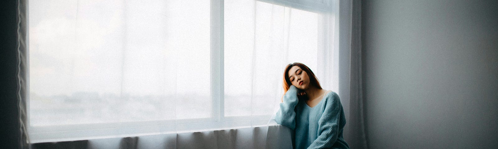 A woman in a blue smock sits on the floor beside a curtained window, her elbow resting on the window sill.