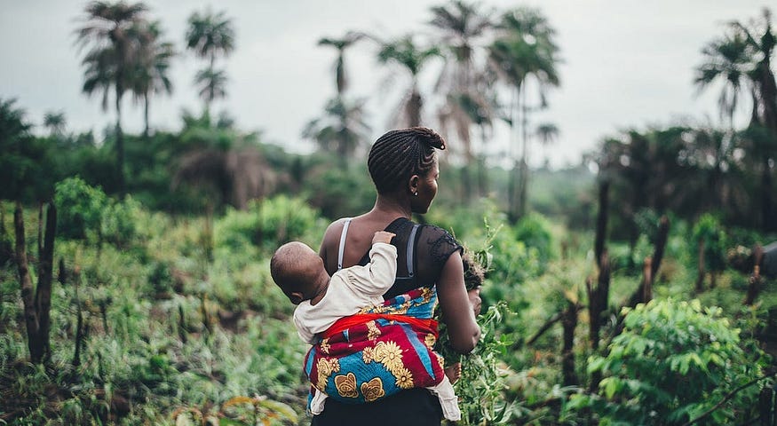 A photo of an African woman carrying her son on her back while she walk through her farm. The son is curious looking around him.