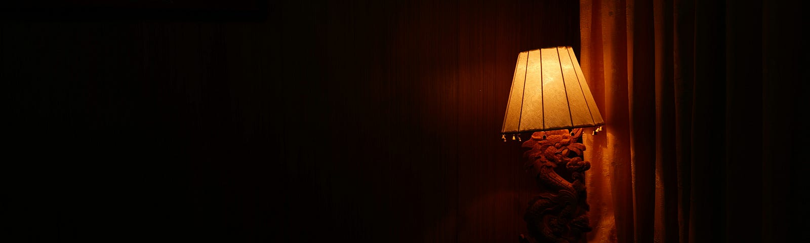 A darkened bedroom illuminated only by a low-powered bedside lamp, casting a soft, warm glow in the otherwise shadowed room