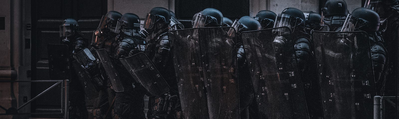 A line of armored police officers hold their ground at a protest. Their gear is spray painted black, obscuring all of their identifying features, and they hold shields and clubs at the ready.