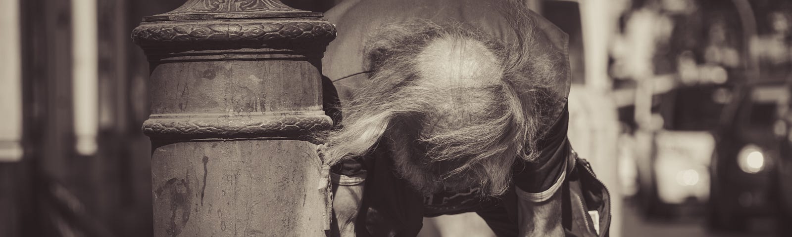 Elderly man with long hair but balding, washing his hands from a fire hydrant that is trickling a small stream of water.