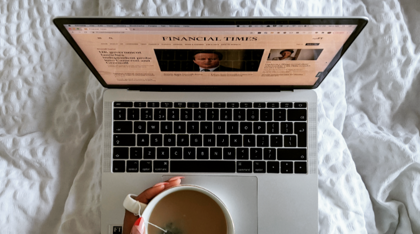 Laptop showing the Financial Times homepage on a bed with a cup of tea in hand and funky red and pink nails