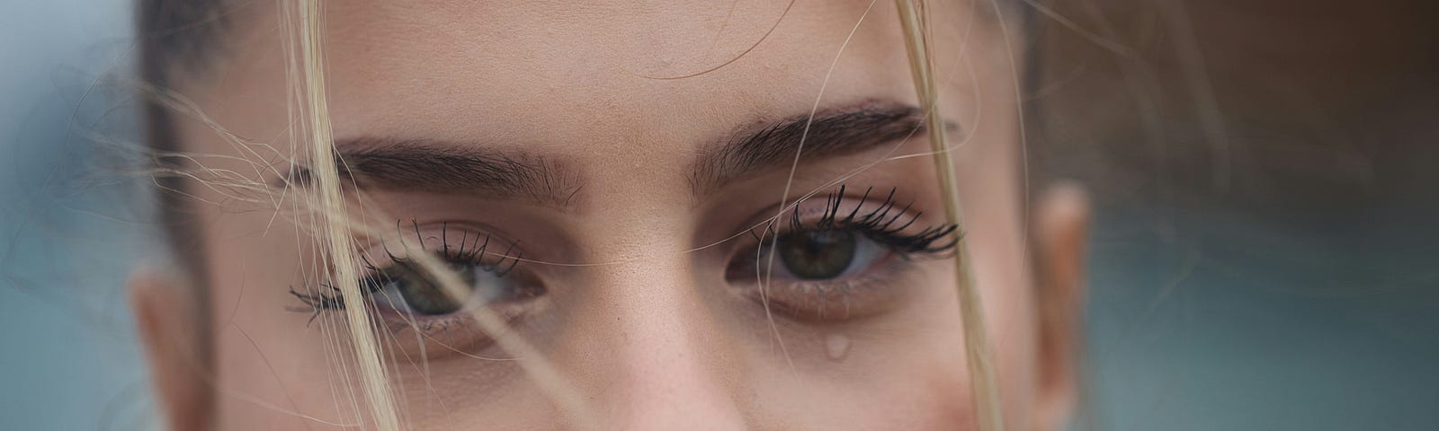 A pretty woman with strong eyebrows and long eyelashes sheds a tear. The look on her face is pained and she has hot pink lipstick on.