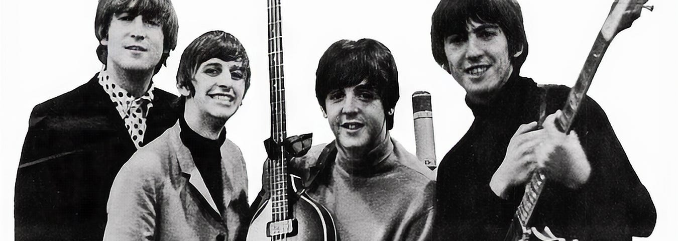 The Beatles band photo in black & white; band members left to right: John Lennon, Ringo Starr, Paul McCartney holding his bass guitar with the headstock facing the sky, microphone between McCartney and George Harrison with his guitar pointed top right