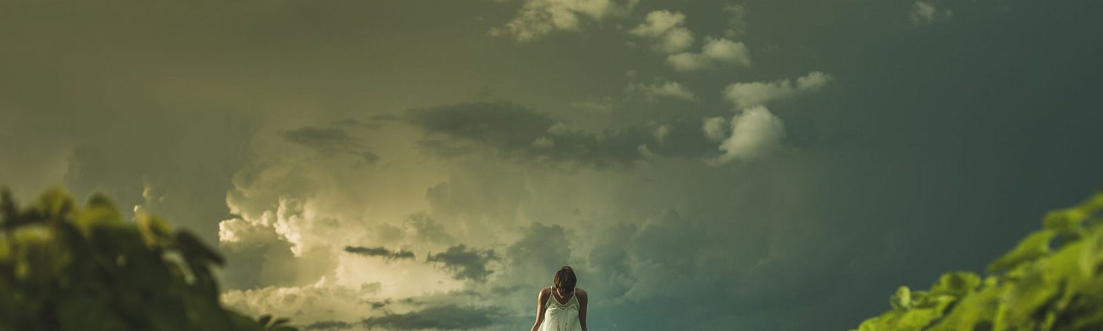 Picture of femme person in white dress with their head looking down, standing in greenery, swirling clouds behind them