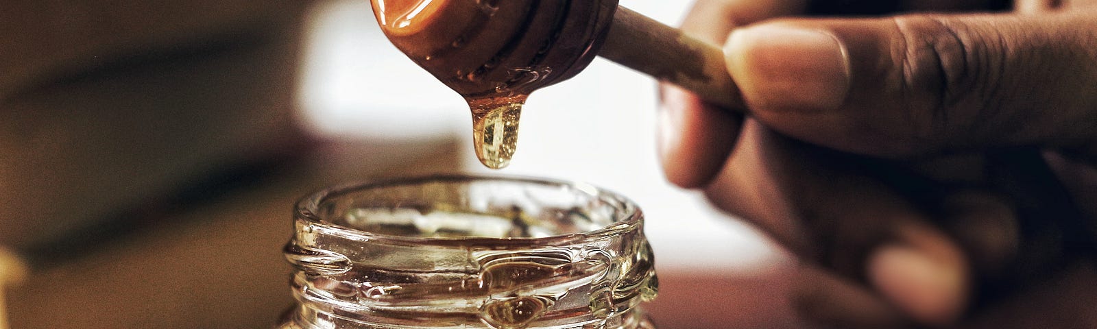 A hand lifts a honey dipper over a jar of honey. A single golden drop of honey is suspended from the dipper.