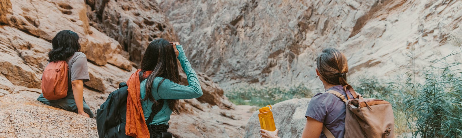 Three women, backs to the camera, sitting on rocks. They are wearing backpacks and looking up towards a rocky passage.