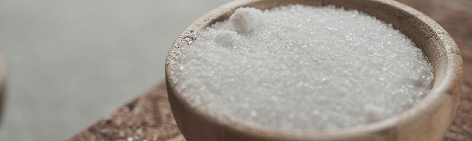 White sugar in a small pale wooden bowl, on a grainy wood table top.