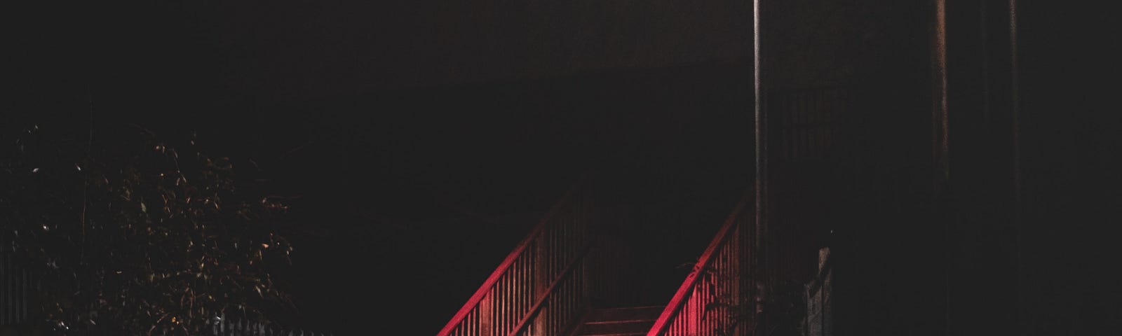 A stairway with red railings, illuminated by a street lamp at night while it’s raining.