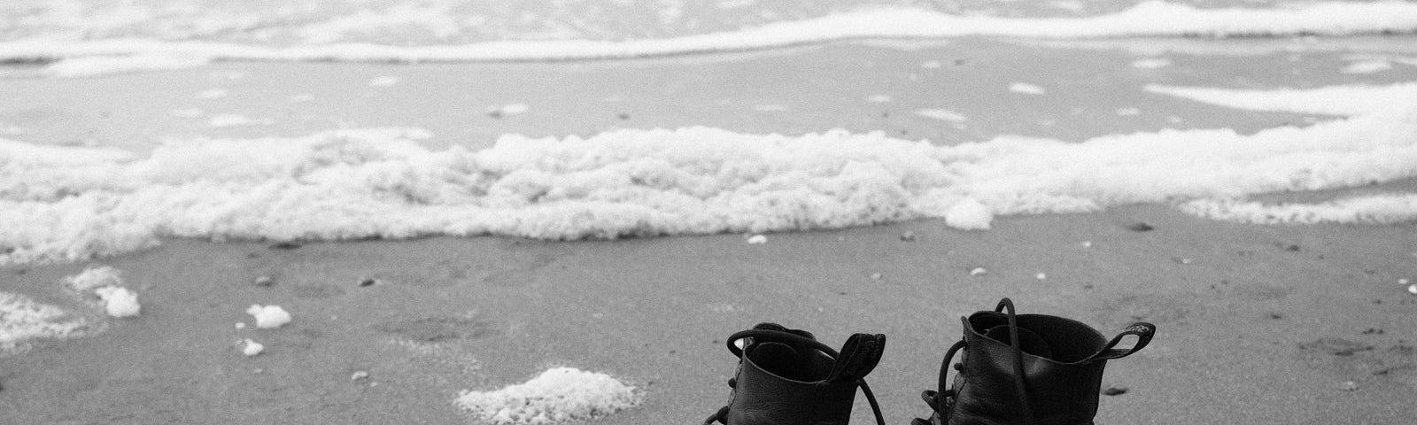 A black-and-white photograph of an empty pair of black shoe-boots at the edge of calm ocean water at the shore