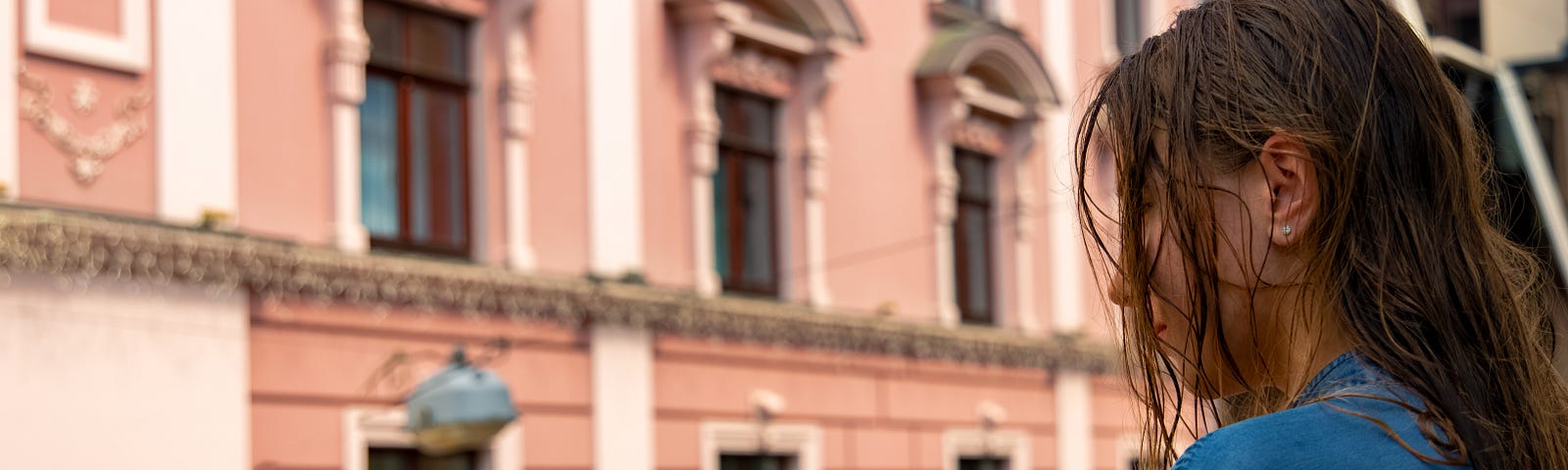 An attractive looking girl, see from a shoulder view, looks out over a balcony to a nice stone building at the other side of the street.