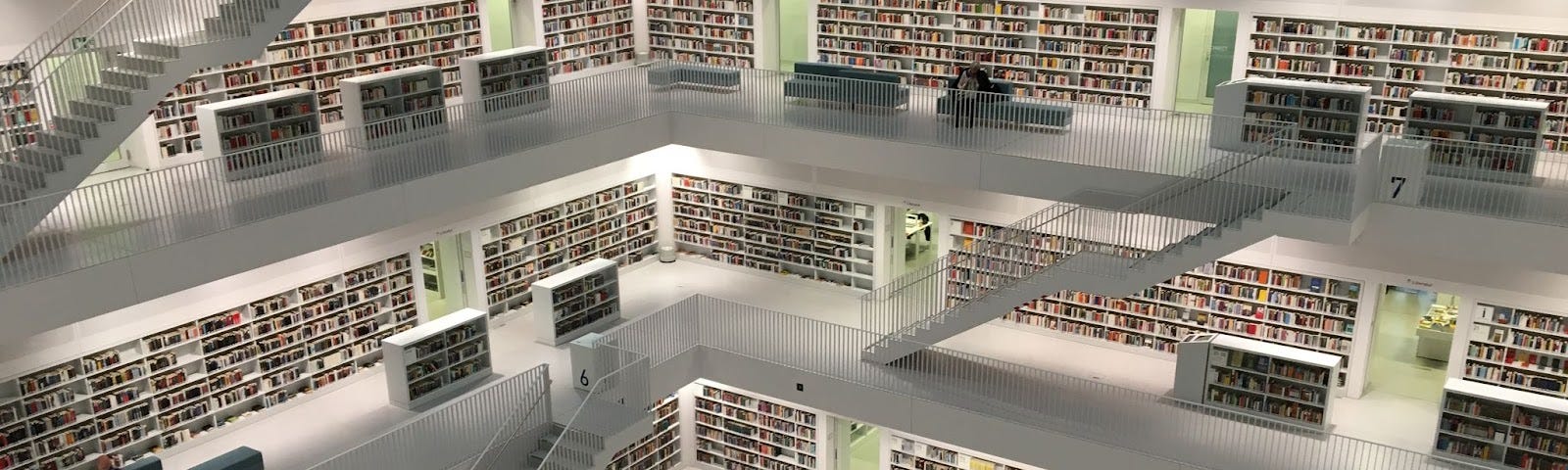 A library with five levels connected by white staircases. Blue couches are on each level, some have readers sitting on them with a book. Bookcases are white filled with rows and rows of books.