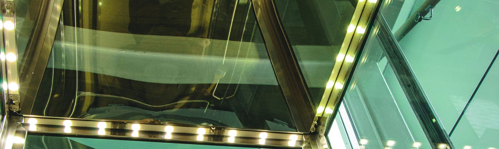 A woman, dressed in stylish leather with round glasses, looks up in an elevator at a reflection of herself on the ceiling.