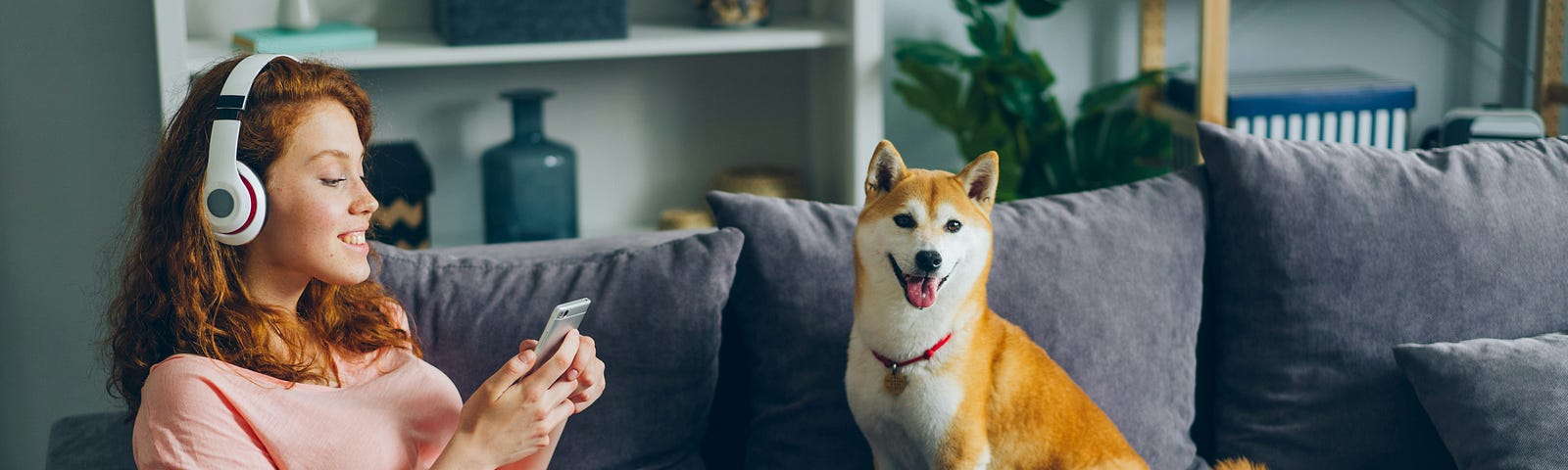 A lady lies on a sofa with her dog. She is looking at her phone and wearing headphones.