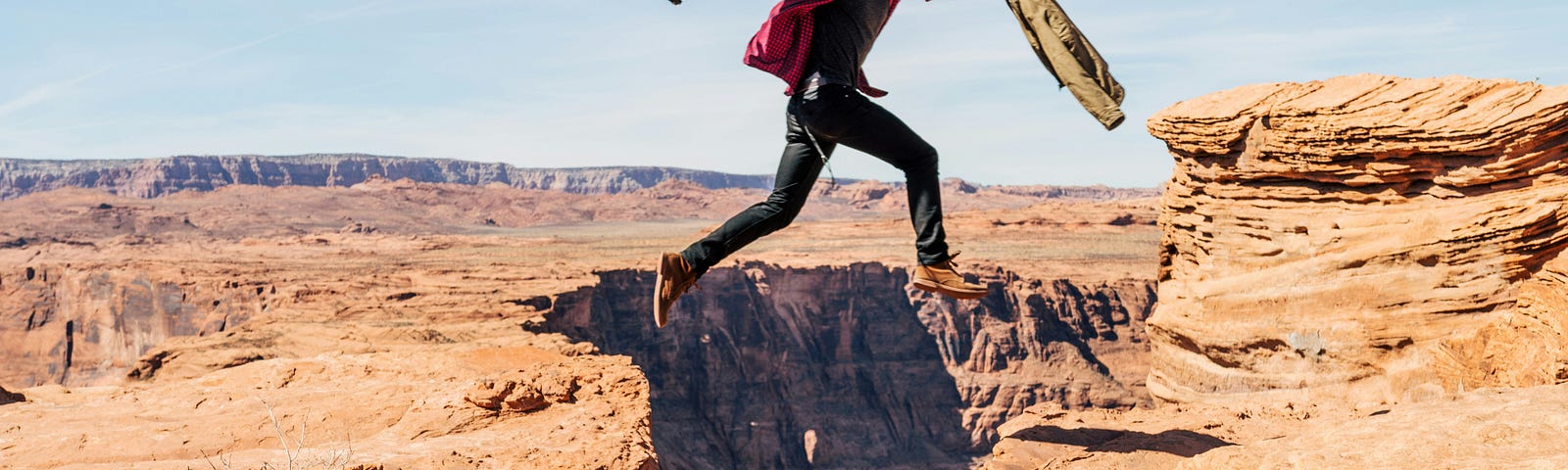 A man holding his jacket and a camera is suspended mid-jump between two cliffs on a sunny day.