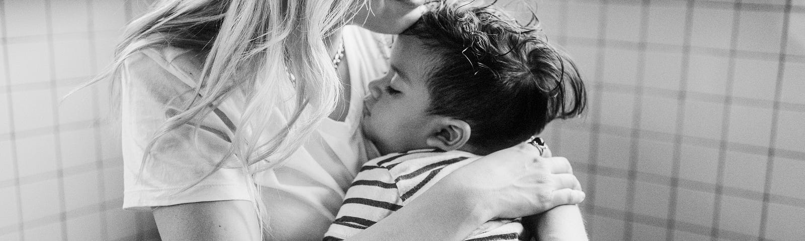 Black and white photo of a mother with long blond hair cradling a sleeping toddler boy in her lap, with his head against her shoulder.