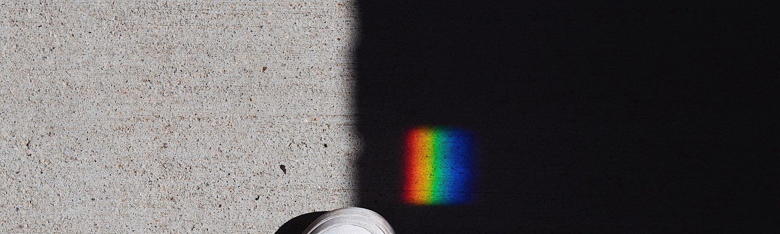 A white shoe on a pavement bisected by a dark, black shadow. To the left is light-gray pavement. To the right is the shadow. Above the show is a small square patch of rainbow light.