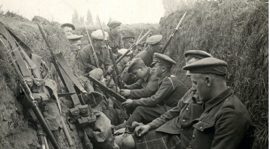 A black and white photograph of soldiers sitting silently in a trench and wearing kilts, army coats, caps, long socks and lace-up boots. Rifles and kitbags rest on the trench walls and some are held by soldiers, while a dog sits near a soldier at the front of the picture.