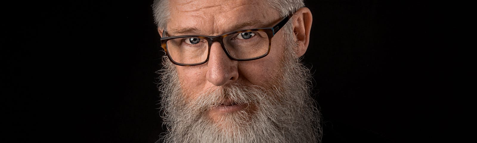Man with white hair and beard wearing glasses and a black shirt on a black background