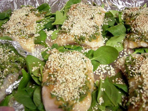 Uncooked catfish fillets covered with herbs, spices, and sesame seeds on beds of spinach leaves on a foil covered baking sheet.
