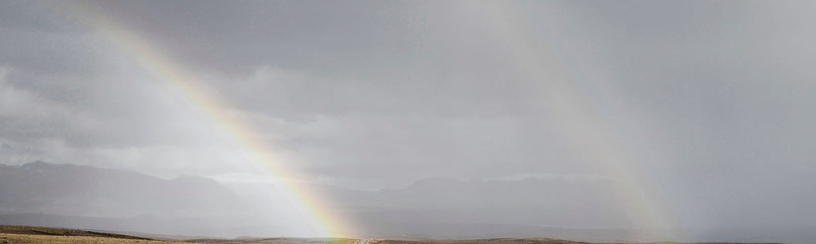 Photo of a road winding through open moorland with a grey sky and two rainbows in the distance.