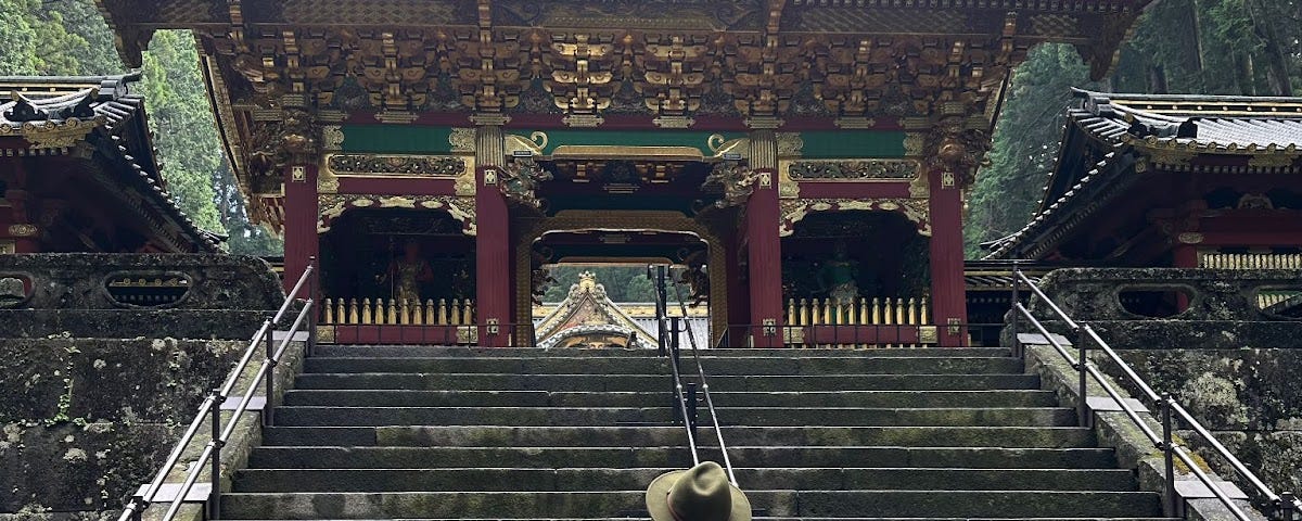Man photographed from behind in green t-shirt, white shorts and with a small black backpack and green side bag. Green felt fedora. Looking off at the steps leading to a Japanese Buddhist temple.