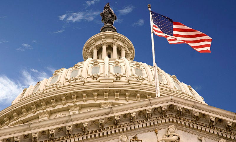 Image of the United States Capitol Building Dome, from a bottom-up perspective. Dome is white against a blue sky, with a big American flag waving in the wind.